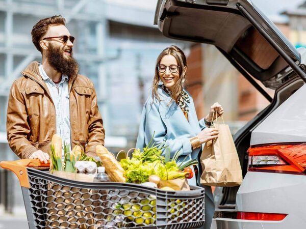 Auf dem Bild sind zwei Personen mit einem gefüllten Einkaufswagen zu sehen, die ihre Einkäufe in ein Auto laden.