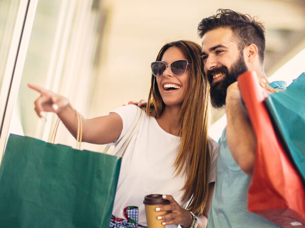 Frau und Mann mit Einkaufstaschen vor einem Schaufenster, lachen glücklich während eines begeisternden Shoppingerlebnisses.