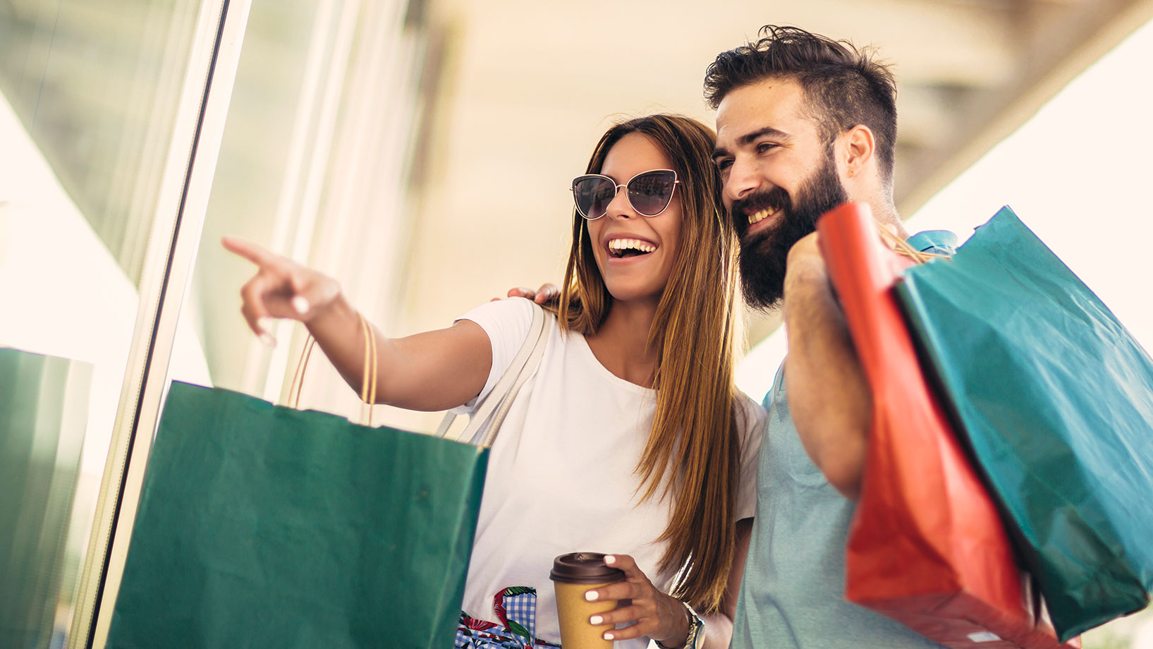 Frau und Mann mit Einkaufstaschen vor einem Schaufenster, lachen glücklich während eines begeisternden Shoppingerlebnisses.