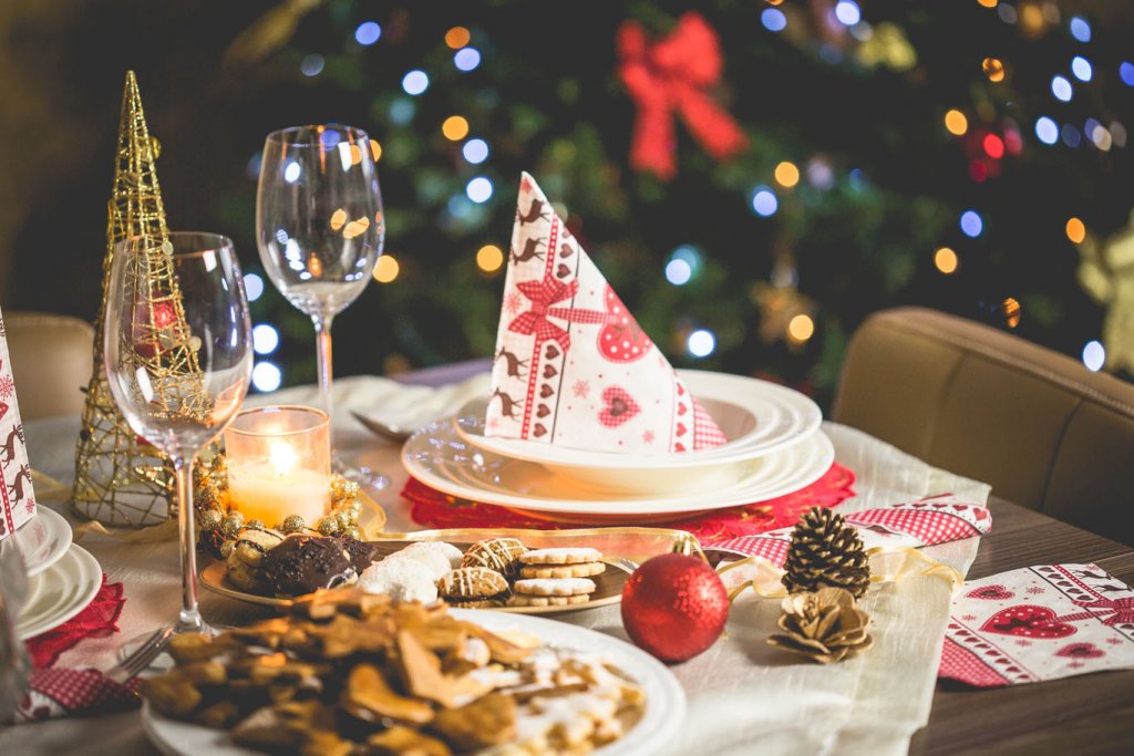 Es ist ein weihnachtlich gedeckter Tisch zu sehen. In der Mitte befindet sich ein Teller mit aufgestellter Serviette, links daneben stehen zwei Weingläser, vorne liegen Weihnachtsplätzchen auf einem Teller, daneben liegt Dekoration und im Hintergrund ist steht ein Weihnachtsbaum.