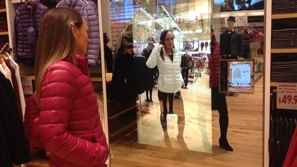Eine Frau mit einer roten Jacke steht vor einem Magic Mirror im Geschäft, der eine ganzheitlich personalisierte Customer Journey ermöglicht.