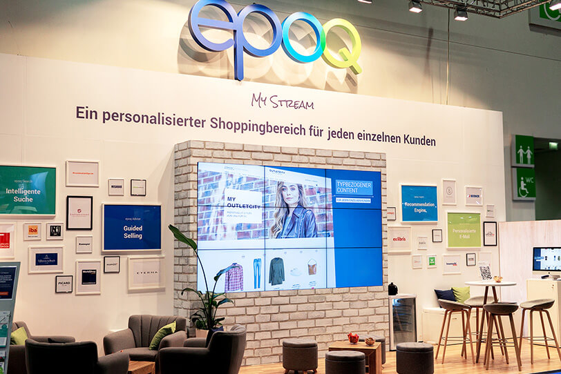 Das Bild zeigt den Stand von epoq auf der DMEXCO 2019. Der Stand ist wie ein Wohnzimmer aufgebaut mit verschiedenen Sitzmöglichkeiten und einem großen Bildschirm.