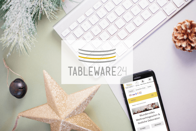 Das Bild zeigt einen weihnachtlich dekorierten Schreibtisch. Darauf liegt ein Smartphone, auf dem die Weihnachtsmarkt-Themenseite des Online Shops von Tableware24 zu sehen ist.